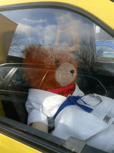 A teddy bear dressed in a white outfit with a blue belt and glasses sitting in the driver's seat of a yellow car, visible through the window.