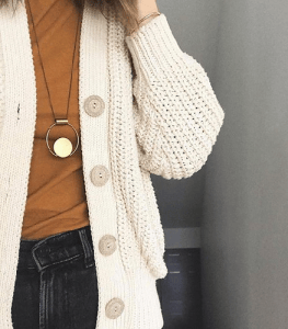 Close-up of a person's arm wearing a cream-colored cardigan with buttons, over a brown top, paired with dark jeans and a circular necklace.